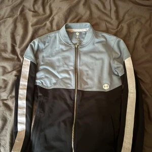 Under Armour ziptröja blå och svart - Snygg ziptröja från Under Armour i ljusblått och svart med vita detaljer längs ärmarna. Tröjan har hel dragkedja framtill, ståkrage och logga på bröstet. Perfekt för träning eller chill. Materialet är polyester och passformen är normal.