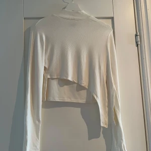 Vit croppad långärmad topp från SHEIN - Säljer en vit croppad långärmad topp från SHEIN i ribbad polyester. Toppen har asymmetrisk nederkant och långa ärmar med slitsdetalj. 