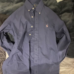 Mörkblå skjorta från Ralph Lauren - Snygg mörkblå skjorta från Ralph Lauren med klassisk button-down krage och vita knappar. Skjortan har lång ärm och den ikoniska rosa broderade loggan på bröstet. Tillverkad i bomull och har en normal passform. Storlek S. Gott skick 