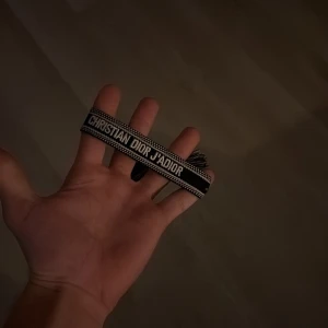 Christian Dior J'Adior armband svart - Snyggt svart armband från Christian Dior med texten 'J'Adior' broderad i vitt. Armbandet har en vävd design med fransar i ändarna och är perfekt för dig som vill ha en exklusiv accessoar med tydlig logga.