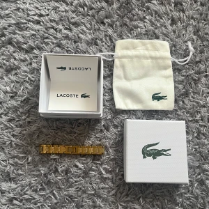 Guld Lacoste Armband - Snyggt armband från Lacoste i guldigt metallutförande med länkdesign och präglade detaljer. Armbandet har den klassiska Lacoste-loggan på en av länkarna och kommer med original ask och tygpåse. Perfekt accessoar för att lyfta din outfit. Den är 17cm och funkar även att ändra i storlek genom att ta ut olika bitar. Byten funkar