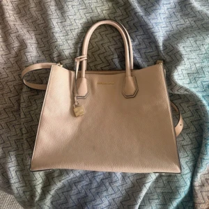 Beige handväska från Michael Kors - Snygg och stilren beige handväska från Michael Kors i skinn. Väskan har två handtag, axelrem och guldfärgade detaljer. Invändigt finns flera fack och dragkedja. Den är köpt i Danmark för många år sen, ej använd mycket.