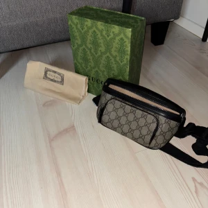 Gucci magväska - Säljer en snygg magväska från Gucci med klassiskt GG-mönster i beige och brunt. Väskan har svarta skinn- och textildetaljer, justerbart band och dragkedja framtill. Perfekt för att bära det viktigaste och ge din outfit en lyxig touch. Skriv vid frågor Elelr intressen 