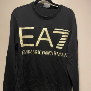 Svart EA7 Emporio Armani tröja - Svart långärmad tröja från EA7 Emporio Armani med stor guldig logga framtill. Tröjan har rund halsringning och är tillverkad i mjuk bomull. Perfekt för dig som gillar stilrena och exklusiva plagg med tydlig branding.