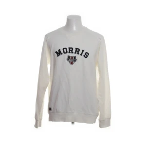 Vit sweatshirt från Morris - Vit sweatshirt från Morris Gents med svart broderad text och emblem på bröstet. Klassisk rund halsringning och långa ärmar. Tröjan är i mjuk bomull och har ribbade muddar vid ärmslut och nederkant. Perfekt för en clean och sportig look.