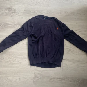 Mörkblå tröja från Polo Ralph Lauren - Mörkblå långärmad tröja från Polo Ralph Lauren med klassisk rund halsringning och den ikoniska röda logotypen broderad på bröstet. Tröjan är i mjuk bomull och har ribbade muddar vid ärmslut och nederkant. Perfekt för en stilren look.