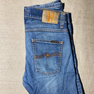 Blå Nudie Jeans - Snygga blå jeans från Nudie Jeans med klassisk orange söm och vågig brodyr på bakfickan. Modellen är W30 L30 GRIM TRIM med slitning och hål på ena knät för en cool, avslappnad look. Jeansen har fem fickor och är tillverkade i mjukt denimtyg. W30L30 grimtrim  vid frågor pris kan alltid diskuteras‼️♟️