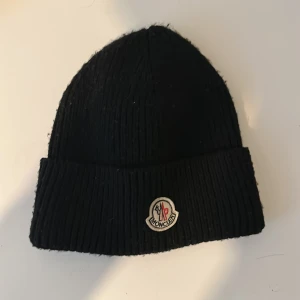 Svart ribbstickad mössa från Moncler - Svart ribbstickad mössa från Moncler med klassisk uppvikt kant och broderad logotyp framtill. Perfekt för kalla dagar och ger en clean streetstyle-look. Materialet är mjukt och värmande, och mössan har en enkel, tidlös design.