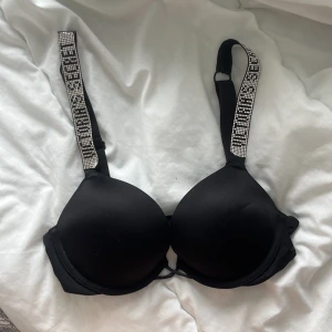 Svart push-up bh Victoria's Secret - Svart push-up bh från Victoria's Secret med breda axelband dekorerade med glittrande stenar och logotyp. Bh:n har vadderade kupor och justerbara axelband för perfekt passform. Står ingen storlek men skulle säga att det är 70A/B.