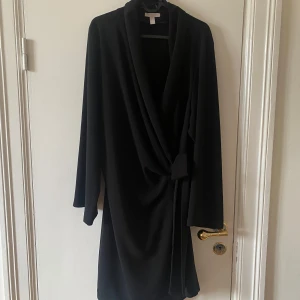 Svart kavajklänning från H&M, XL - Svart kavajklänning från H&M i storlek XL. Klänningen har långa ärmar, omlottkrage och knytband i midjan. Ovan knä längd.