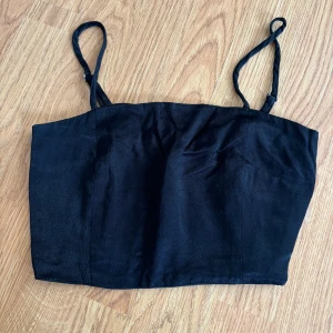 Svart linnetopp från Zara i satin - Snygg svart linnetopp från Zara i glansigt satinliknande material. Toppen har smala, justerbara axelband och smockad rygg för extra komfort. Enkel och stilren design som är lätt att matcha med olika outfits.