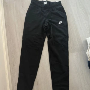 Svarta mjukisbyxor från Nike, stl S - Svarta mjukisbyxor från Nike med klassisk vit logga på låret. Byxorna har resår i midjan med snörning och är tillverkade i mjukt material som passar perfekt för chill eller träning. Snygg och enkel design som är lätt att matcha med hoodies eller t-shirts. Storlek s 