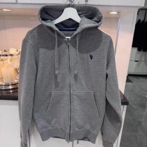 Grå hoodie från U.S. Polo Assn, S - Grå hoodie från U.S. Polo Assn med dragkedja, huva och snörning. Broderad marinblå logga på bröstet och två stora fickor framtill. Skön och stilren, perfekt för chill dagar eller när du vill ha en enkel look. storlek S och helt oanvänd!