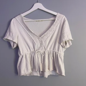 Vit blus med spets från Pull&Bear - Supersöt vit blus från Pull&Bear med v-ringning, broderade spetsdetaljer och knyt framtill. Blusen har korta ärmar och volang nedtill för en romantisk vibe. Perfekt till jeans eller kjol för en fräsch look.