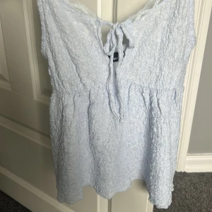 Ljusblå spetsdetaljerad linnetopp Gina Tricot - Supersöt ljusblå linnetopp från Gina Tricot med tunna axelband och spetsdetaljer längs kanterna. Toppen har en knytning framtill och en lätt skrynklig struktur som ger en chill vibe. Perfekt för varma dagar och enkel att styla med jeans eller kjol.