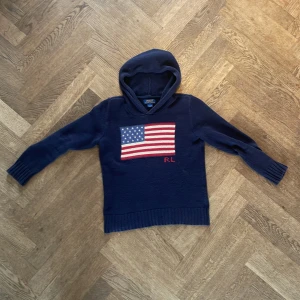 Ralph lauren hoodie/sweater - Mörkblå stickad hoodie från Polo Ralph Lauren med amerikansk flagga och RL-broderi på bröstet. Tröjan har huva, långa ärmar och ribbade muddar. Perfekt för dig som gillar klassisk och sportig stil. Perfekt inför hösten och vintern🍂❄️ 172 på bild