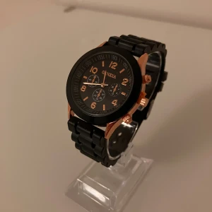 Svart och guldig klocka från Geneva - Stilren klocka från Geneva med svart urtavla och armband. Klockan har detaljer i guld, inklusive siffror, visare och knappar. Boetten är rund och armbandet är länkformat i svart. Perfekt accessoar för dig som gillar en klassisk men modern look.                                                ✅Frakt inom 24 timmar 📦.                                                      ✅Nytt Batteri 🔋.