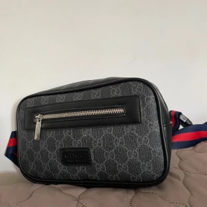 Svart axelväska  - Snygg svart axelväska med klassiskt GG-mönster i grått. Väskan har en silverfärgad dragkedja framtill och bred axelrem i blått och rött. Tillverkad i canvas och skinn med detaljer i svart läder. Perfekt för dig som vill ha en stilren och trendig accessoar.