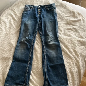 Blå bootcut jeans från Zara, stl 164 - Snygga blå jeans från Zara med bootcut-modell och hög midja. Jeansen har flera silverfärgade knappar framtill och klassiska fickor. Tillverkade i jeansmaterial med coola slitningar och kontrastsömmar. Perfekta för dig som gillar en avslappnad men trendig look.