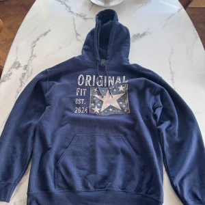 Mörkblå hoodie från Scndi med tryck - Säljer en mörkblå hoodie från Scndi med stort tryck framtill där det står 'ORIGINAL FIT EST. 2024' och en stjärna. Tröjan har huva med snörning och en stor magficka. Perfekt för chill dagar eller när du vill ha en avslappnad stil. Lite ludd på tröjan men går bort på en tvät. Skriv vid minsta funderingen!