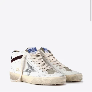 Golden Goose Mid Star sneakers med glitter - Golden Goose Mid Star sneakers i vitt skinn med grå mockadetaljer och en glittrig silverstjärna på sidan. Skorna har snörning, perforerade detaljer och en mörkröd rand på sidan. Snygg kontrast med blå hälflik och beige sula. Perfekta för dig som gillar unika detaljer.