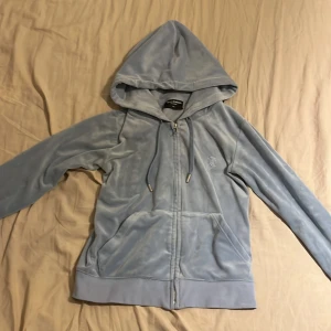 Ljusblå velour hoodie Juicy Couture XS - Supermjuk ljusblå hoodie från Juicy Couture i velour med dragkedja, huva och snörning. Klassisk logga broderad på bröstet och två fickor framtill. Perfekt för en chill och trendig look.