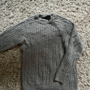 Grå kabelstickad tröja från Next - Säljer en grå kabelstickad tröja från Next i 100% bomull. Tröjan har rund halsringning, ribbade muddar och lång ärm. Perfekt för dig som gillar klassisk stil med en twist. Passar dig som är ca 165-175 cm lång.