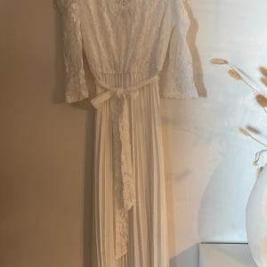 Vit långklänning i spets med bälte - Superfin vit långklänning med spets upptill, långa ärmar och plisserad kjol. Klänningen har ett knytbälte i midjan och medföljer ett glittrigt bälte med silverdetaljer. Perfekt för speciella tillfällen!