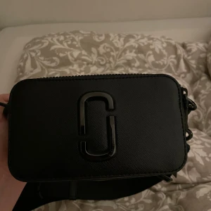 Svart axelväska från Marc Jacobs - Snygg svart axelväska från Marc Jacobs i stilren boxig modell. Väskan är tillverkad i strukturerat skinn och har en tydlig logga framtill samt dragkedja upptill. Perfekt storlek för det viktigaste och en riktigt trendig accessoar🌷 super bra skick, saknas en dragflik. 