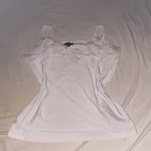 Vit linnetopp med spetsdetaljer - Säljer en vit linnetopp från New Yorker med tunna axelband och vacker spets längs urringningen. Toppen är ärmlös och har en feminin, enkel design som passar perfekt till jeans eller kjol. Materialet känns mjukt och lätt mot huden.