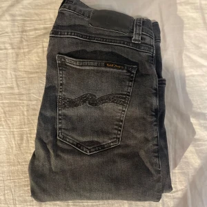 Svarta nudie jeans - Snygga svarta jeans från Nudie Jeans med klassisk femficksdesign och diskret logga på bakfickan. Jeansen har en mörk tvätt och smal passform som sitter tajt längs benen. Perfekta för dig som gillar en stilren och modern look.