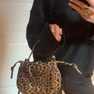 Leopardmönstrad axelväska med snörning - Cool axelväska i leopardmönster med justerbar axelrem och snygga snörningsdetaljer på sidorna. Väskan är i bruna och svarta toner och har en rundad form. Perfekt för dig som vill sticka ut med en trendig accessoar.