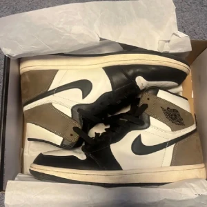 Jordan 1 Mocha - Nike Air Jordan 1 Retro High OG i färgen Dark Mocha, svart och vit. Klassisk high-top siluett med svart swoosh, snörning och rund tå. Ovandel i skinn och mocka för en lyxig känsla. Perfekt för dig som vill sticka ut med en ikonisk sneaker. Perfekt för hösten!