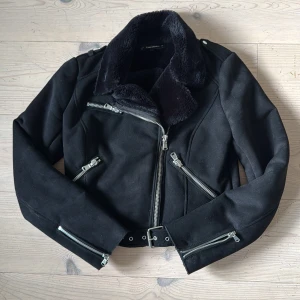 Svart teddy bikerjacka från Zara - Snygg svart teddyjacka från Zara med bred fuskpäls-krage, flera silvriga dragkedjor och bälte i midjan. Jackan har en cool bikerstil och är fodrad med mjuk teddy. Perfekt för dig som vill ha en edgy look med extra värme. Mockaliknande material. 