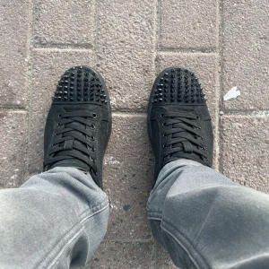 Christian Loubs svarta sneakers med nitar - Svarta sneakers från Christian Loubs med mattsvarta nitar på tån och klassisk röd sula. Skorna har snörning och är tillverkade i mocka och skinn, vilket ger en lyxig känsla. Kommer med originalkartong, dustbag och extra nitar.