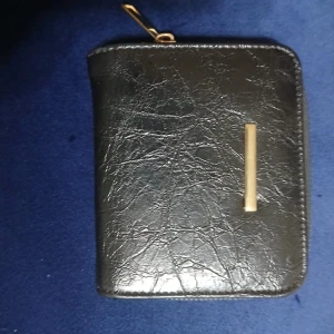 Svart plånbok med gulddetalj - Stilren svart plånbok i skinnimitation med guldfärgad metallstång på framsidan. Plånboken har dragkedja runtom, flera kortfack och ett myntfack med tryckknapp. Perfekt storlek för att få plats i jackfickan eller väskan.