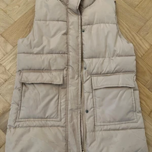 Beige quiltad dunväst med stora fickor - Snygg beige dunväst med quiltad design och två stora fickor framtill. Västen har dragkedja och tryckknappar för extra stilpoäng. Perfekt att slänga över en hoodie eller tröja när det är kyligt ute.
