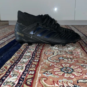 Adidas Predator svarta fotbollsskor - Svarta Adidas Predator fotbollsskor med klassiska tre ränder i mörkblått på sidan. Skorna har snörning, högre stickad strumpkrage och texten 'Predator' i guld på hälen. Tillverkade i syntetmaterial och designade för gräsplaner.