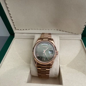 Seiko Daydate Rosé - Säljer en stilren Seiko klocka i roséguld med länkarmband och grön urtavla. Klockan har dag- och datumvisning, romerska siffror och automatisk Miyota-rörelse. Snygg räfflad bezel och transparent baksida som visar urverket.