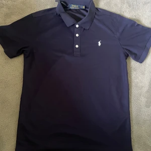 Mörkblå Polo Ralph Lauren t-shirt - Snygg mörkblå t-shirt från Polo Ralph Lauren med klassisk pikékrage och tre vita knappar framtill. Ikoniska loggan broderad på bröstet. Tillverkad i mjuk bomull som känns skön mot huden. Perfekt för dig som gillar stilrena och tidlösa plagg.