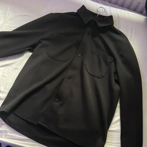Svart overshirt med fickor - Stilren svart overshirt med klassisk krage och två stora bröstfickor med lock. Skjortan har tryckknappar framtill och raka, långa ärmar. Perfekt för lager-på-lager och enkel att matcha med det mesta. Passar otroligt bra på allt o alla. Storlek M