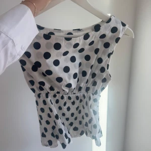 Prickig ärmlös topp från Design by Si - Supersöt vit topp med svarta prickar från Design by Si. Modellen är ärmlös med rund halsringning och har smockad midja som ger en snygg form. Tunt och lätt bomullstyg. Storlek XS/S