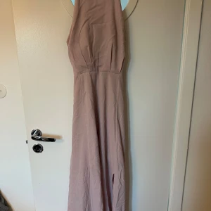 Ljusrosa långklänning Moments NY - Elegant ljusrosa långklänning från Moments New York med spetsdetaljer i ryggen och slits framtill. Klänningen har ärmlös design och stängs med knappar och dragkedja bak. Perfekt för dig som vill sticka ut med en romantisk look.