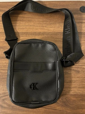 Svart axelväska från Calvin Klein - Snygg svart axelväska från Calvin Klein med justerbar rem och präglad logga på framsidan. Väskan är tillverkad i strukturerat skinnliknande material och har både framficka och bakficka med dragkedja. Perfekt för att bära dina viktigaste grejer.