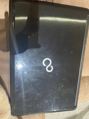 Svart Fujitsu bärbar dator - En svart bärbar dator från Fujitsu med stilren design och rundade kanter. Locket har en blank finish med Fujitsus logotyp i mitten. Chassit är gjort av plast och har en klassisk laptopform.