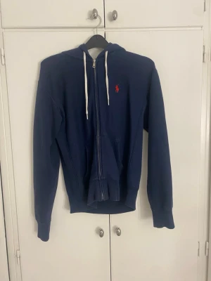 Marinblå hoodie från Ralph Lauren - Snygg marinblå hoodie från Ralph Lauren med dragkedja, vita snören i huvan och den klassiska röda loggan på bröstet. Hoodien har känguruficka framtill och ribbade muddar vid ärmar och nederkant. Perfekt för en chill och stilren look.