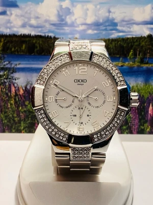 Silverfärgad klocka från OXXO - Stilren och glittrig klocka från OXXO med silverfärgad metallarmband och boett. Urtavlan är vit med stora siffror och flera mindre visare för datum och veckodag. Runt boetten sitter gnistrande stenar som ger extra bling. Perfekt accessoar för dig som gillar att sticka ut.