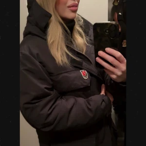 Svart täckjacka från Fjällräven i XS - Snygg svart täckjacka från Fjällräven med klassisk logga på bröstfickan. Jackan har hög krage, huva och dragkedja framtill. Perfekt för kalla dagar och har en stilren, tidlös look.