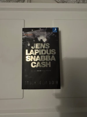 Snabba Cash av Jens Lapidus - Snabba Cash av Jens Lapidus är en svart pocketbok med silverfärgad text på omslaget. Boken tillhör serien Stockholm Noir och handlar om den undre världen i Stockholm. Perfekt för dig som gillar spännande och råa berättelser om brott och maktspel.