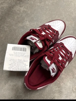 Nike Dunk Low röd/vit sneakers - Nike Dunk Low sneakers i rött och vitt med klassisk låg siluett. Skorna har röd snörning, vit tåbox med perforeringar och röd Nike-logga på plösen och hälen. Ovandelen är i skinn och designen är riktigt clean och tidlös.